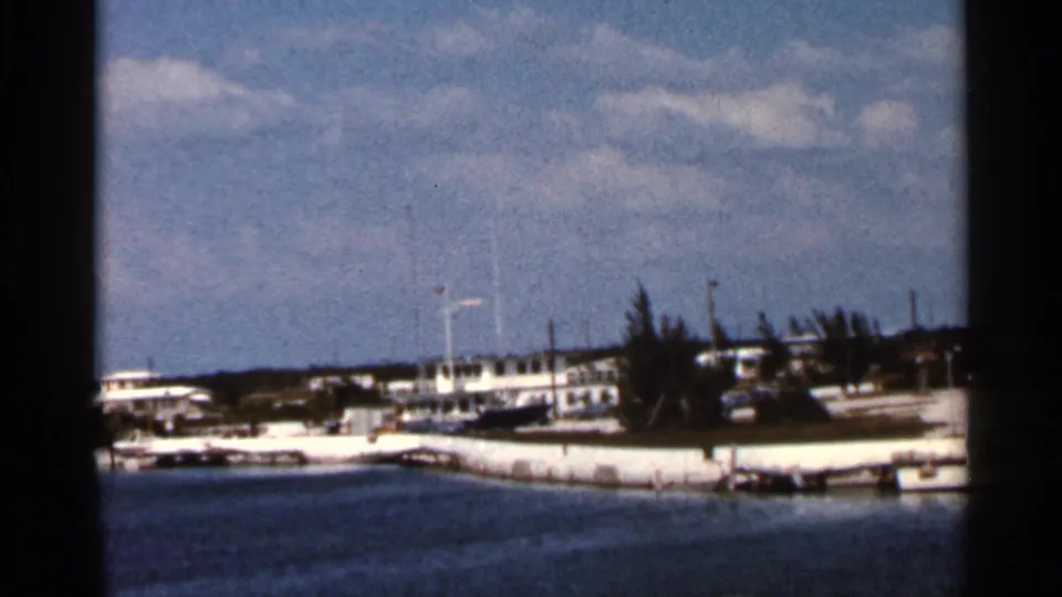Florida shoreline in 1966 home-movie footage, with low white buildings, moored boats, trees, and calm blue water in the foreground.