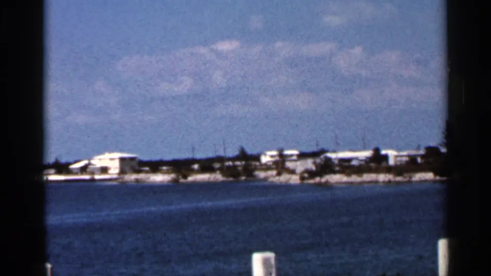 Wider 1966 Florida waterfront frame with open blue water, distant low coastal buildings, trees, shoreline, and a white foreground post.