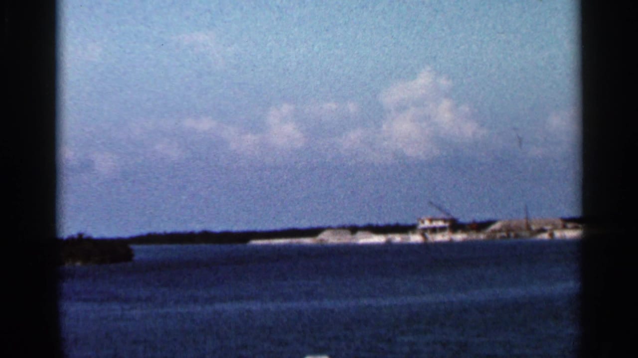 1966 Florida coastal frame with open water in front of a low island and a small structure beside apparent construction equipment.