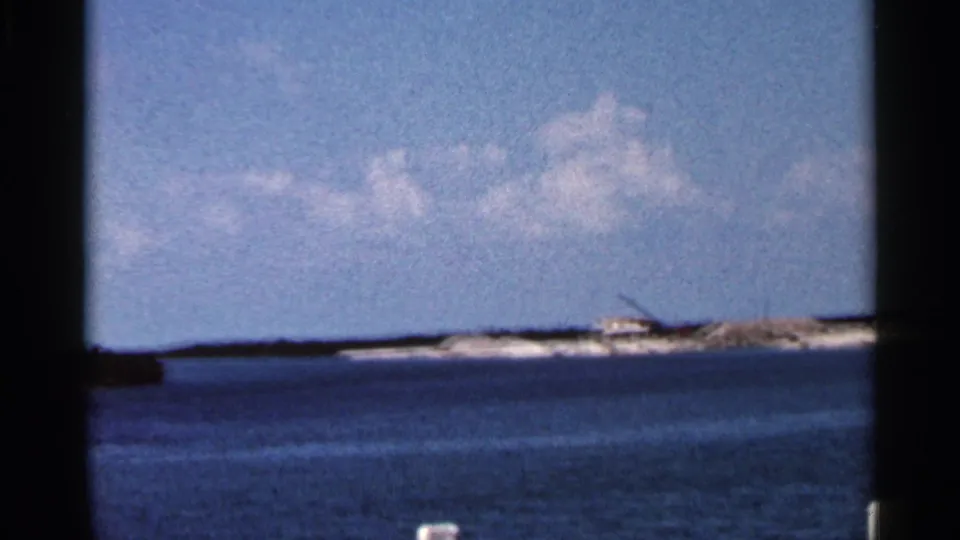1966 Florida home-movie frame looking across blue water to a low island with a small structure and crane-like construction shape.