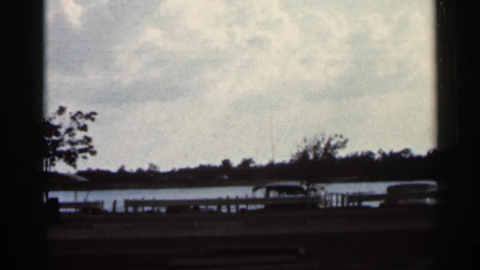 1966 Florida home-movie frame showing a soft view of water, dock railings, shoreline trees, parked cars, and cloudy sky.