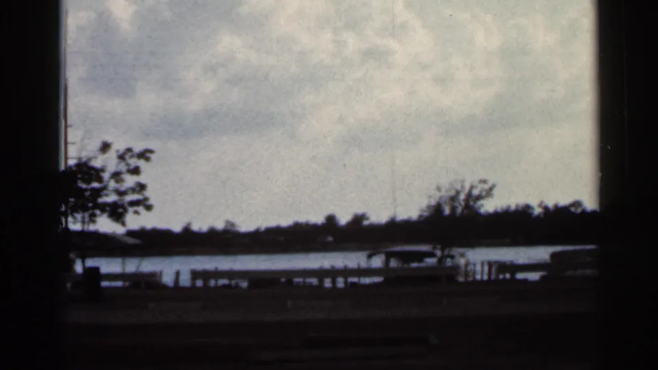 1966 Florida 8mm frame showing a dim waterfront view with water, a distant tree line, low shore structures, and parked cars.