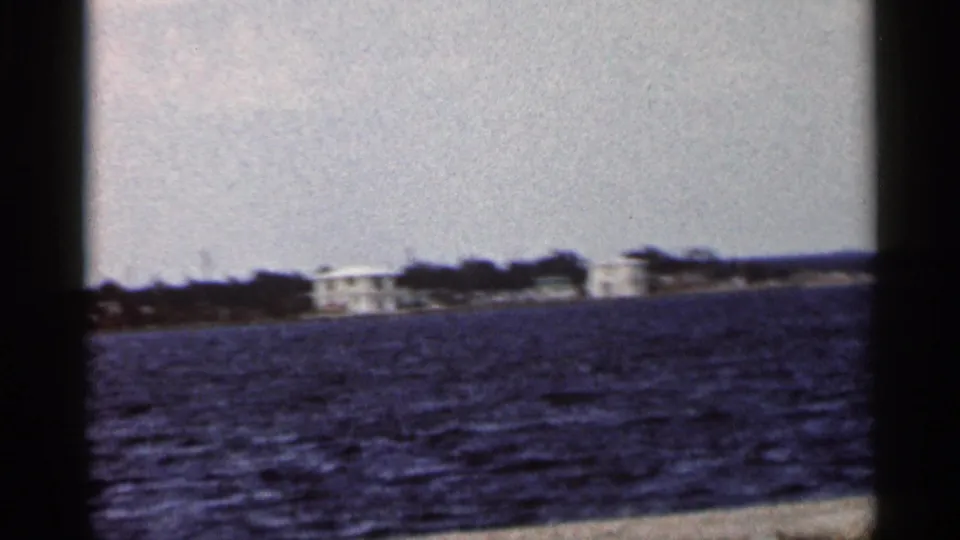 Distant Florida shoreline in 1966 home-movie footage, with white buildings, trees, and open water in soft focus.