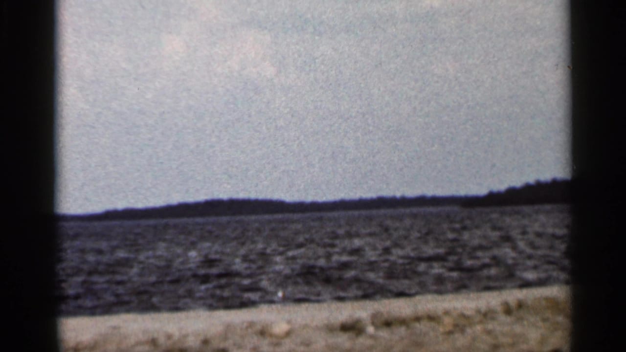 Final 1966 Florida 8mm frame with open water, distant low land, pale sky, and a narrow foreground shoreline edge.