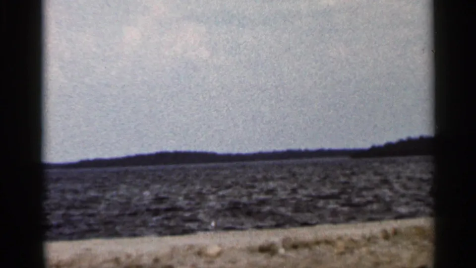 Final 1966 Florida 8mm frame with open water, distant low land, pale sky, and a narrow foreground shoreline edge.