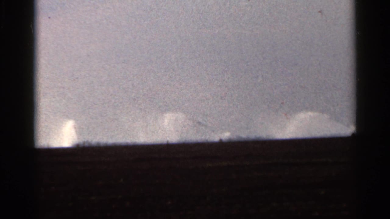1966 Florida home-movie frame with faint irrigation spray and a dark foreground band beneath a muted sky.