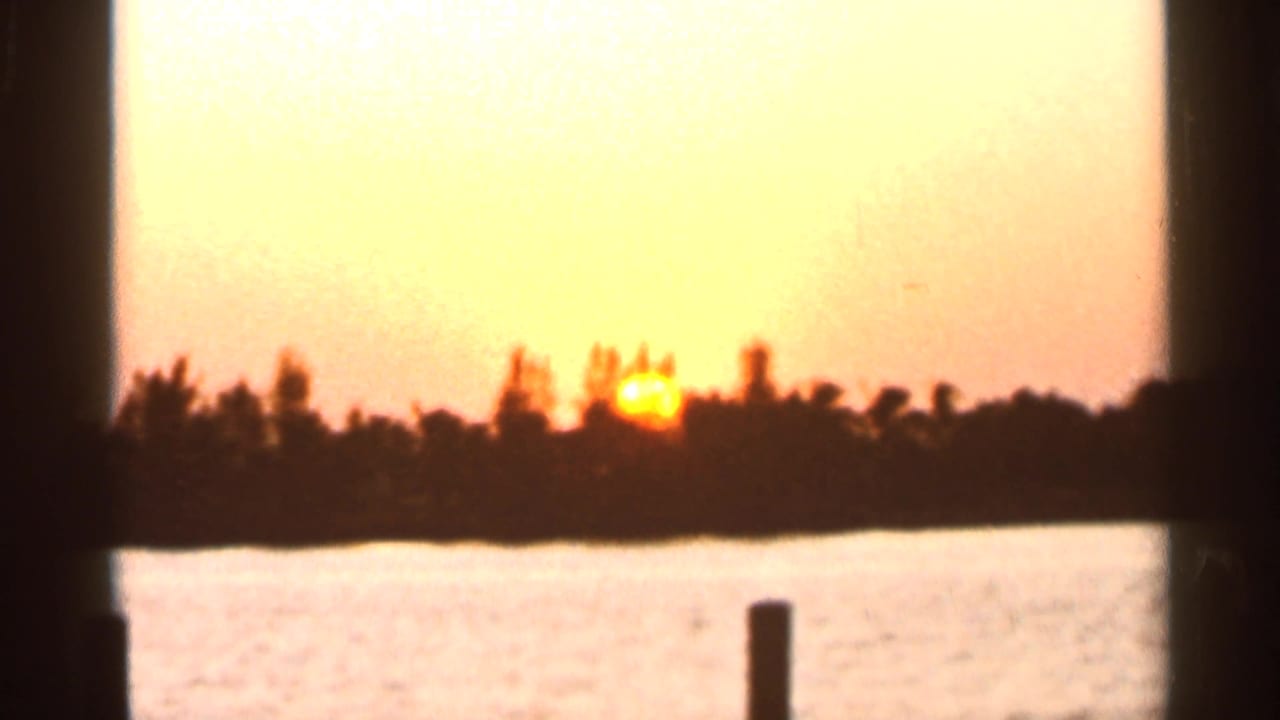1966 Florida home-movie sunset over water, with the sun low behind a silhouetted treeline and ripples in the foreground.