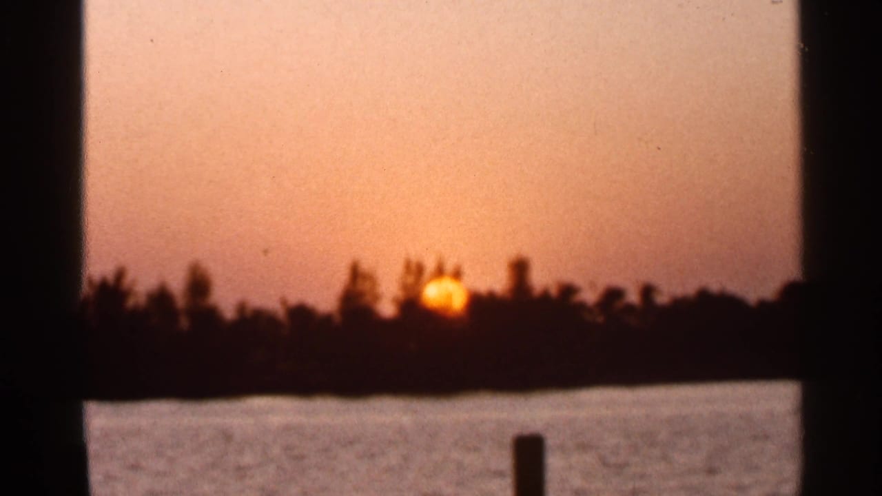1966 Florida 8mm frame showing a low orange sun near the horizon across calm water, with a silhouetted treeline and dark foreground posts.