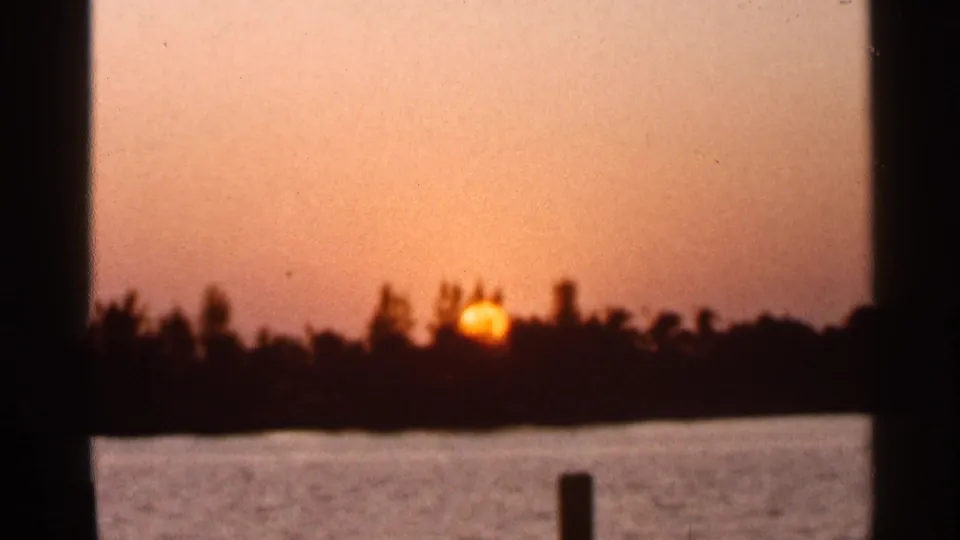 1966 Florida 8mm frame showing a low orange sun near the horizon across calm water, with a silhouetted treeline and dark foreground posts.