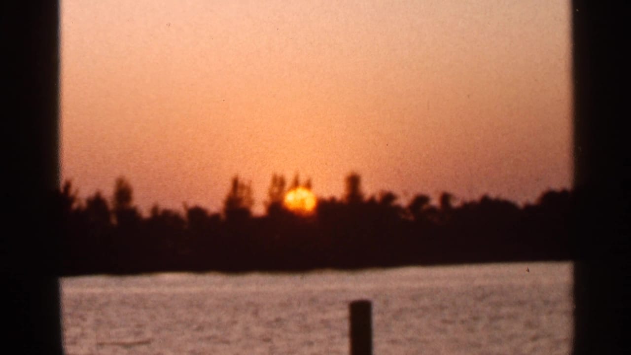1966 Florida home-movie frame of the sun setting beyond a dark treeline across water, seen through vertical foreground shadows.