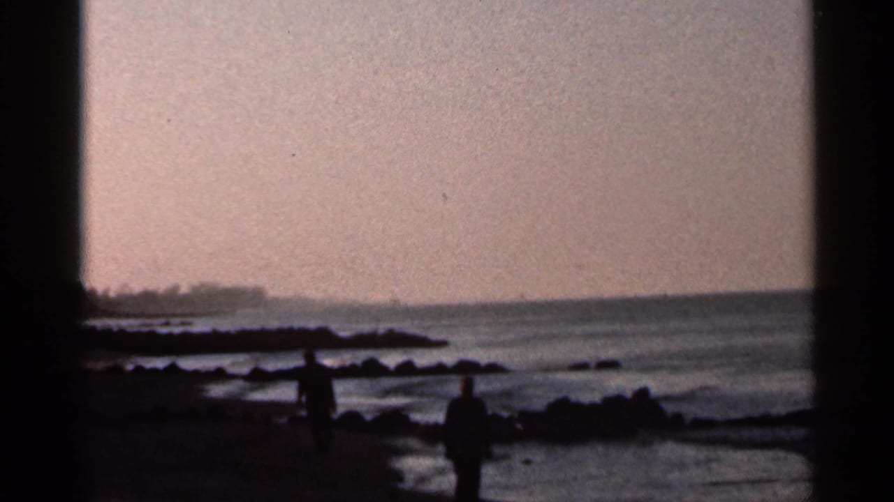1966 Florida shoreline frame showing two distant people walking along wet sand near rocks and calm water.