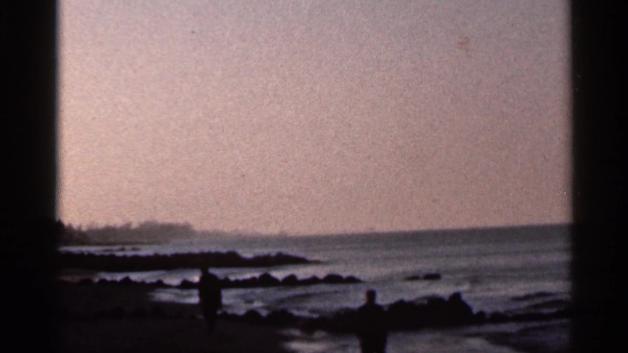 1966 Florida beach scene with distant silhouetted figures near rocks, wet sand, and a calm ocean horizon.