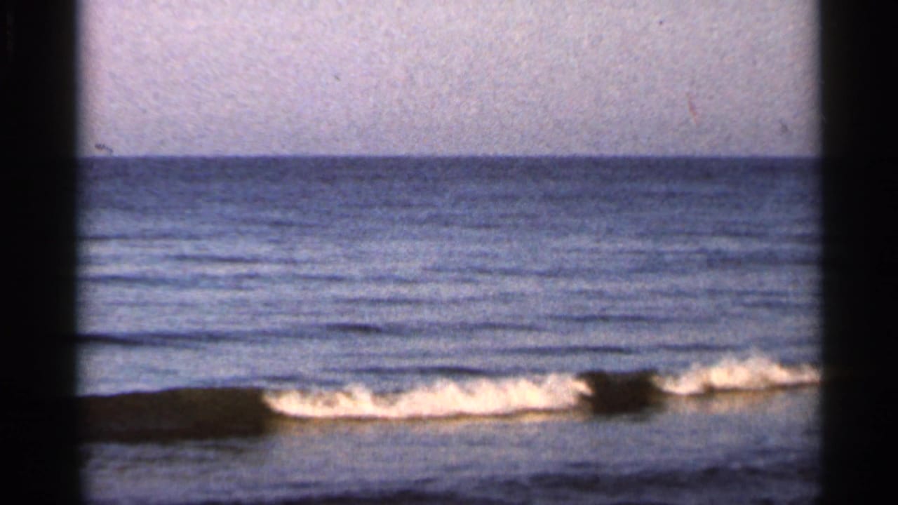 1966 Florida shoreline frame with a small breaking wave, pale sky, soft focus, and visible film grain.