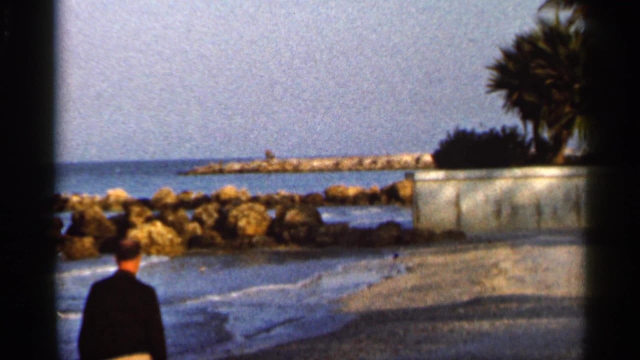 A man in a dark jacket walks along a Florida shoreline near rocks, small waves, a breakwater, and palm trees in 1966 footage.