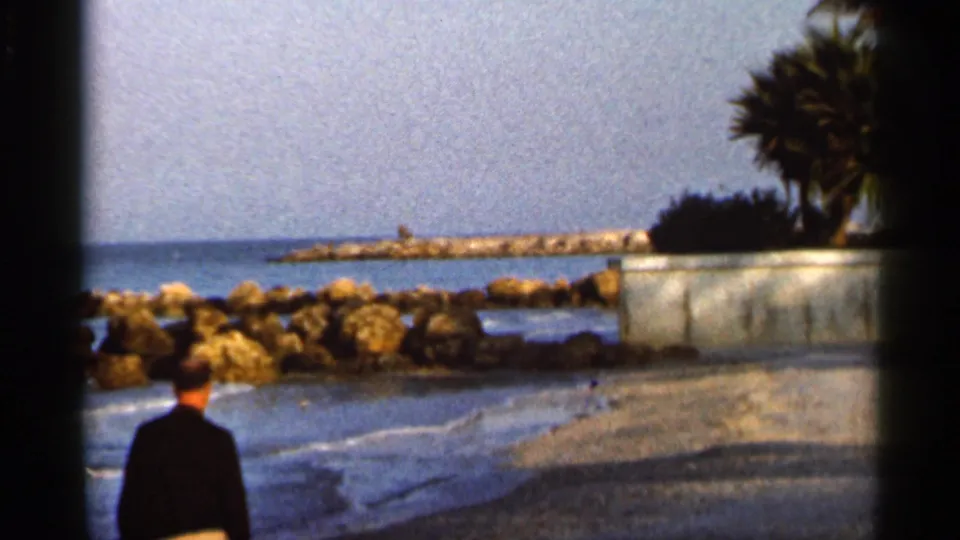 A man in a dark jacket walks along a Florida shoreline near rocks, small waves, a breakwater, and palm trees in 1966 footage.