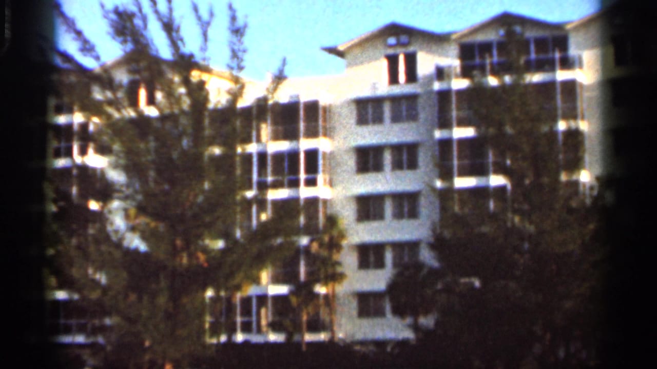 White multi-story resort building partly hidden by trees and palms under a blue sky.