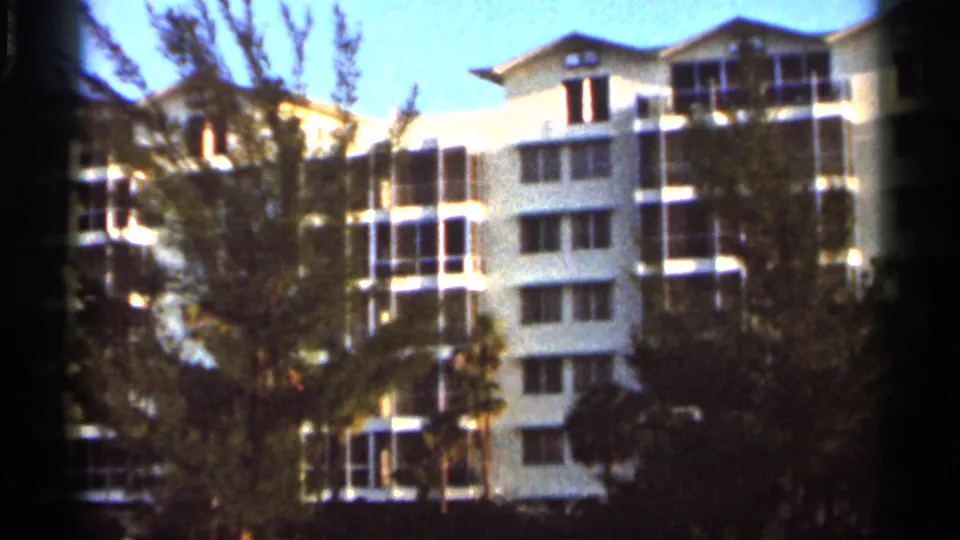 1966 Florida 8mm frame showing a white resort-style building with balconies and dark windows behind palms and leafy trees.