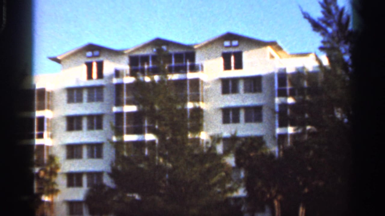 White Florida resort exterior on 8mm film, with upper balconies, rows of windows, palms, and tree cover in the foreground.