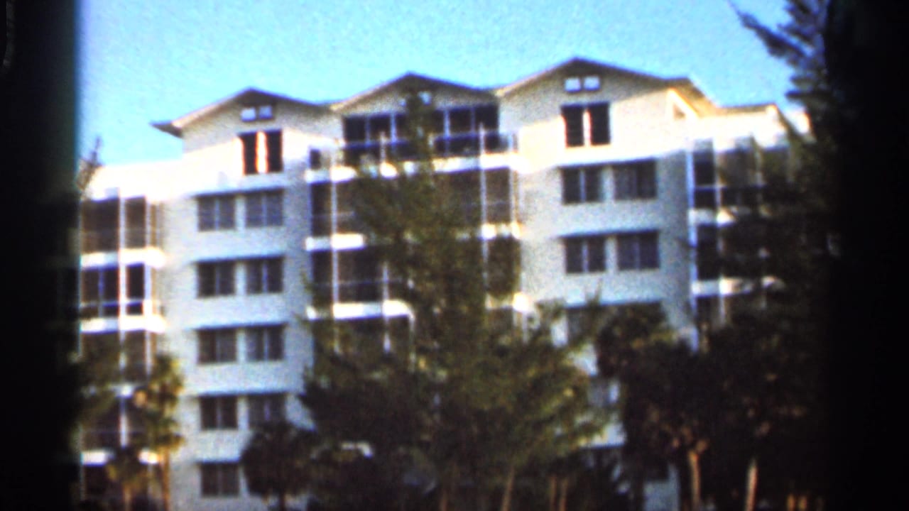 1966 home-movie frame of a white resort building in Florida, partly obscured by trees and palm landscaping.