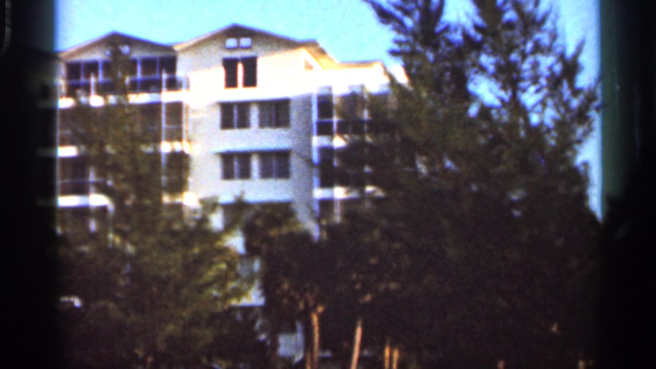 Final 8mm frame showing the Florida resort exterior receding behind darker trees and palm foliage in bright sun.