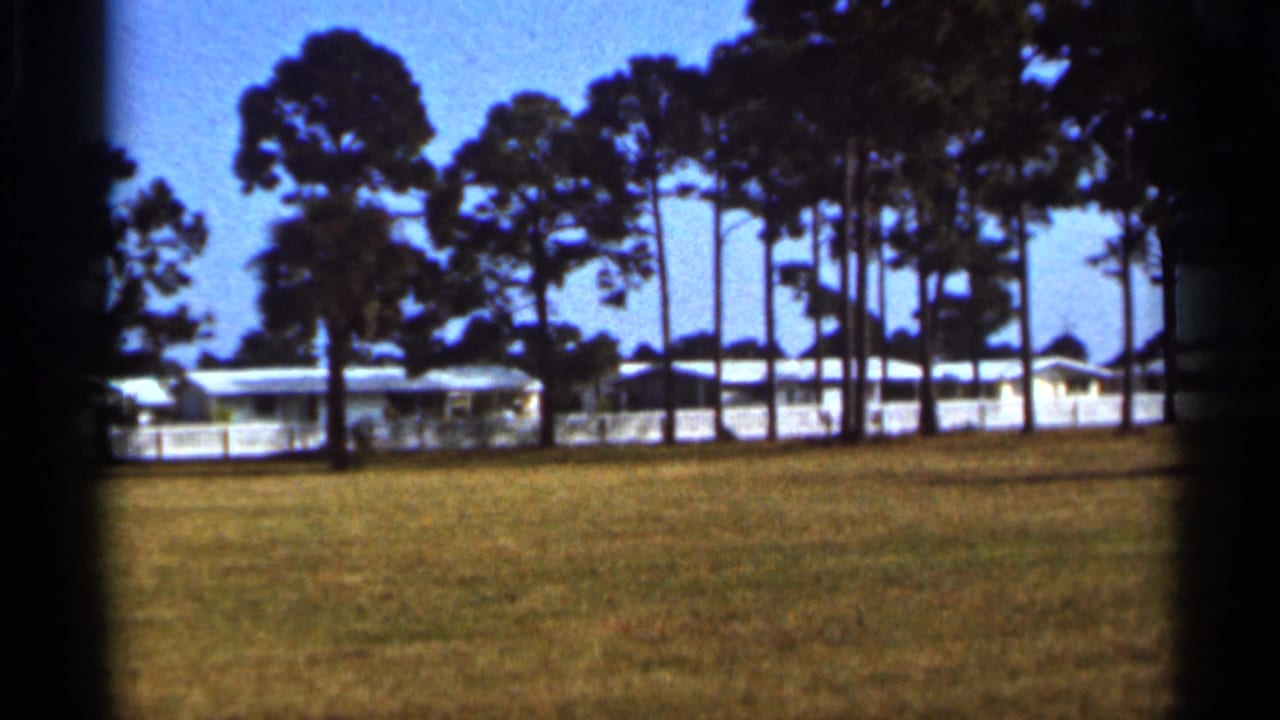 Final 1966 Florida frame showing a pine-lined grassy area with white mobile homes or low buildings under clear blue sky.