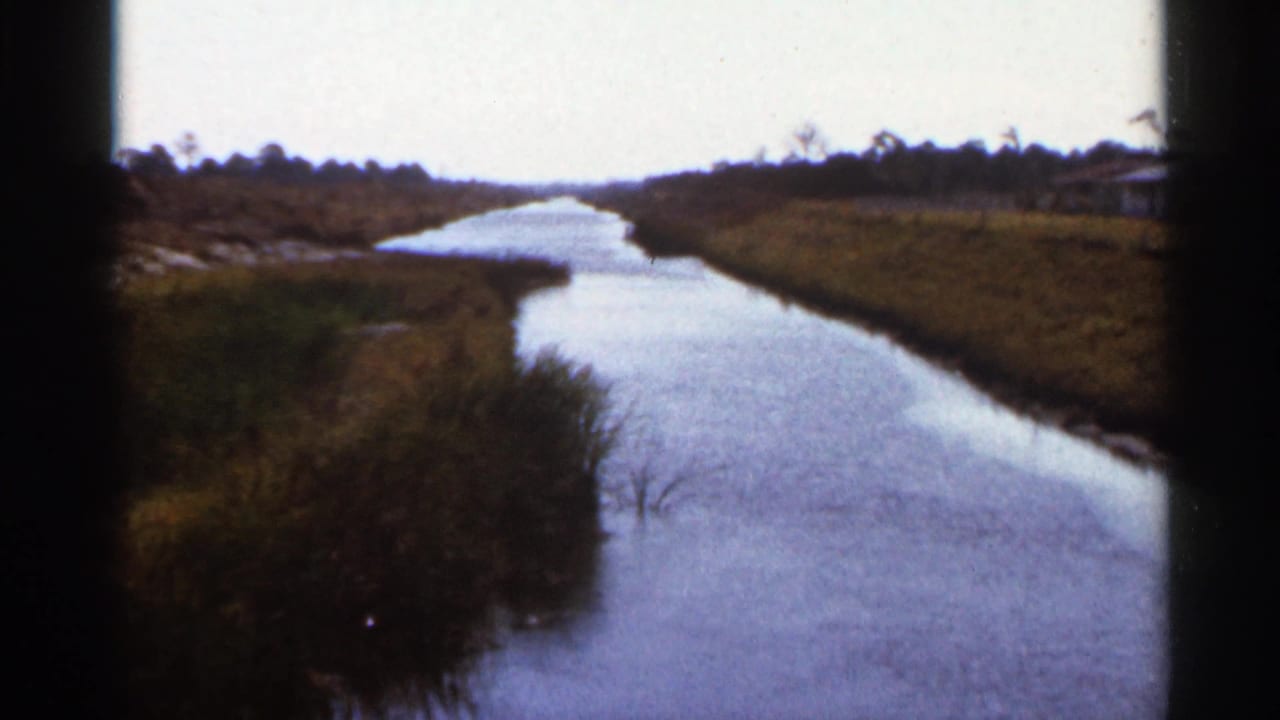 1966 Florida home-movie frame showing a canal curving through marshland with distant low buildings at right.