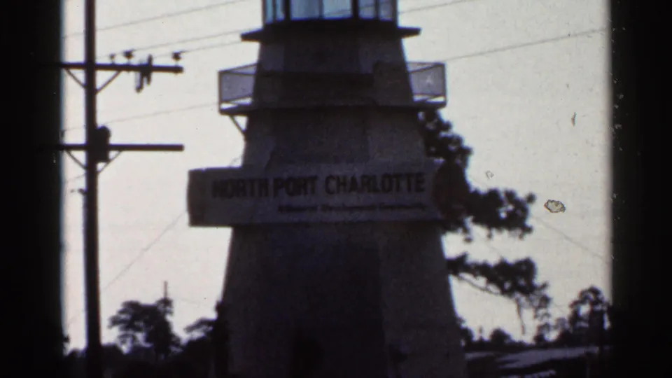 1966 Florida 8mm frame showing a tower sign reading North Port Charlotte, with utility poles, trees, bright sky, and dark frame edges.
