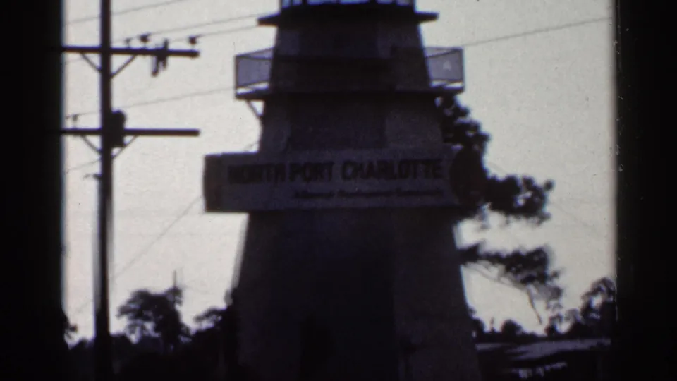 1966 Florida home-movie frame with a soft, dark view of the North Port Charlotte tower sign beside utility lines and trees.