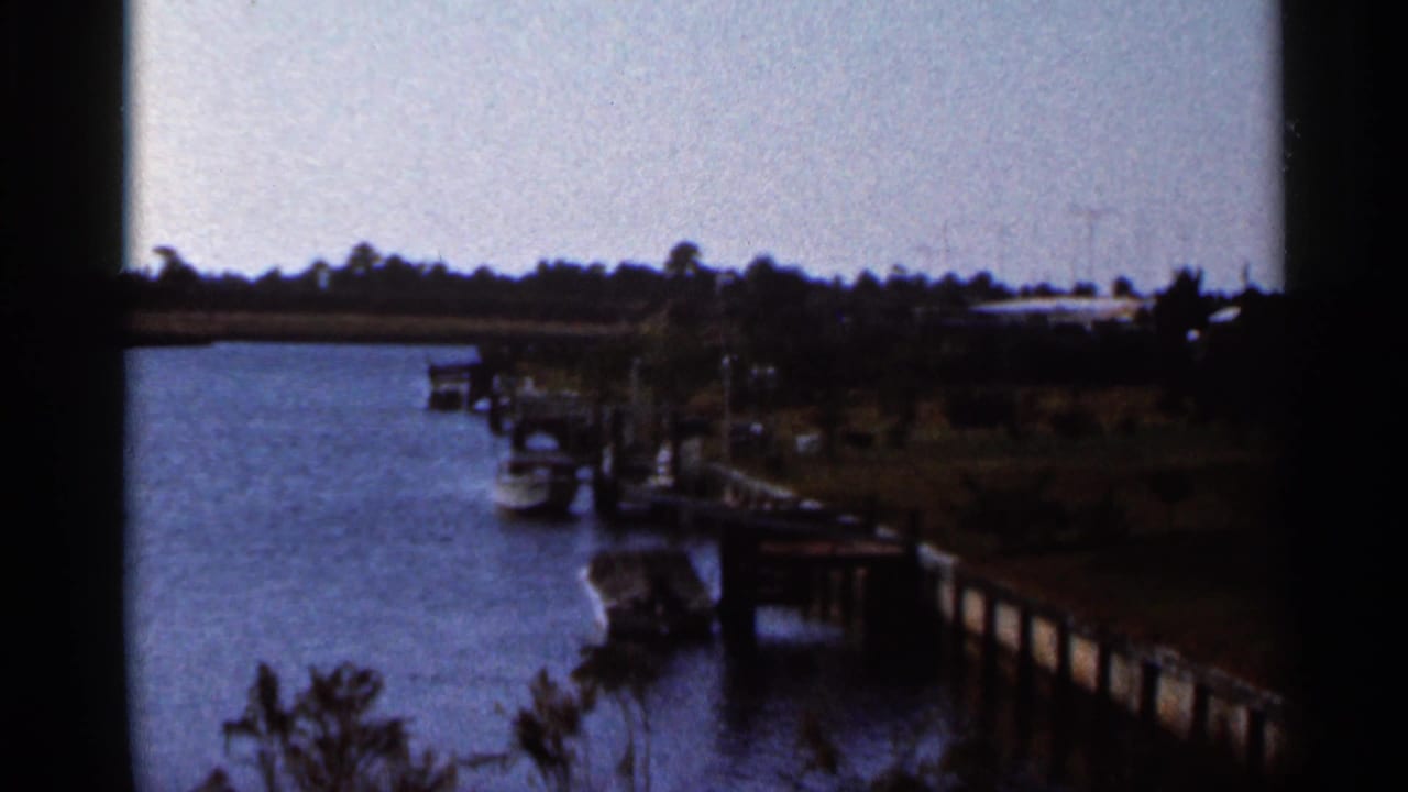1966 Florida 8mm view across a calm river toward docks, moored boats, shoreline trees, and low riverfront structures.