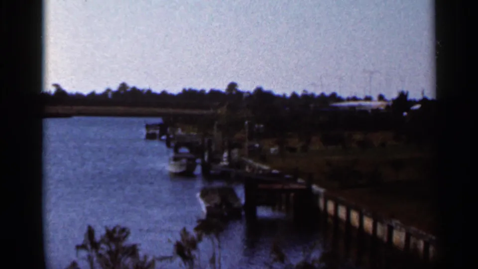 1966 Florida 8mm view across a calm river toward docks, moored boats, shoreline trees, and low riverfront structures.