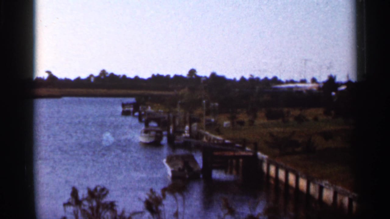 Faded 1966 Florida home-movie frame showing a river marina with docks, small boats, shoreline vegetation, and a pale sky.