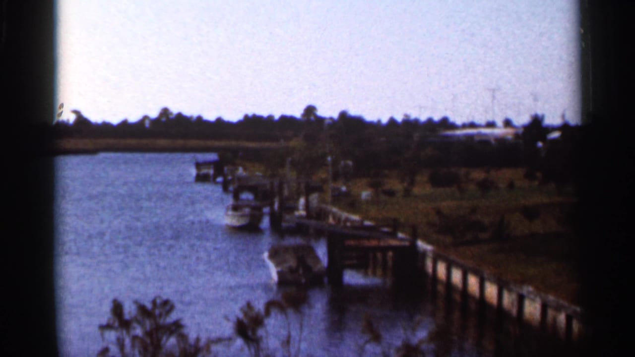 1966 Florida 8mm frame showing a wooden dock, moored boats, blue water, and tropical vegetation along a quiet shoreline.