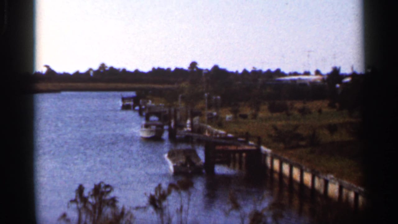1966 Florida home-movie frame of a dock extending along calm water, with small boats and palm trees near the opposite bank.