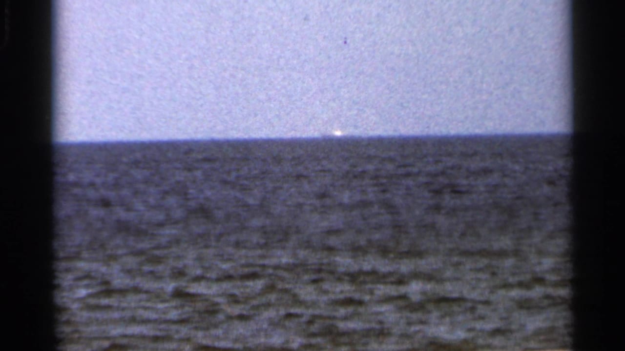 1966 Florida 8mm frame showing textured water, pale sky, and a faint bright object near the distant horizon.