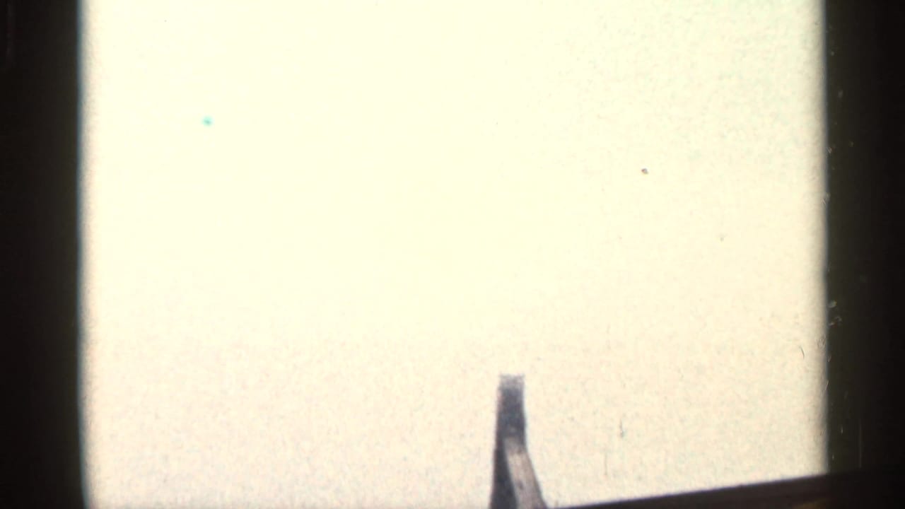 Overexposed 8mm frame or leader area with pale field, dark film edge, and a small dark partial shape near the lower edge.