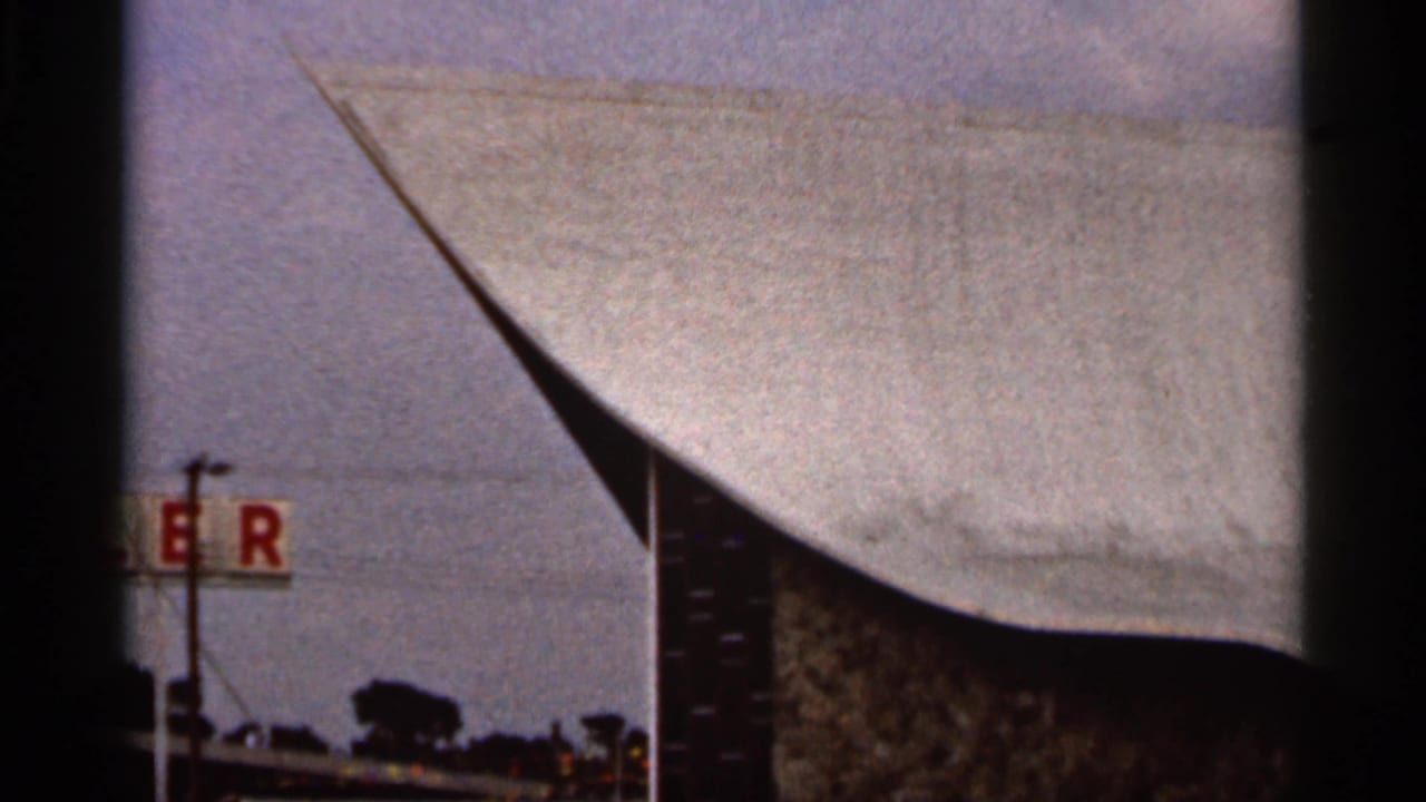 Curved modern church roof with a tall cross and partial red sign at the roadside.