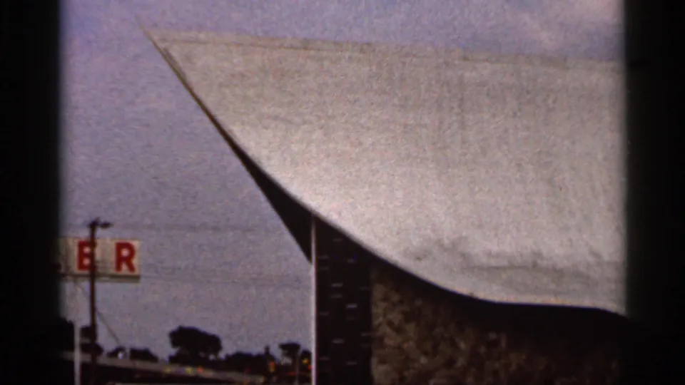 1966 Florida 8mm frame showing a modern church roofline, a tall wooden cross, roadside pole, trees, and a partial red sign.