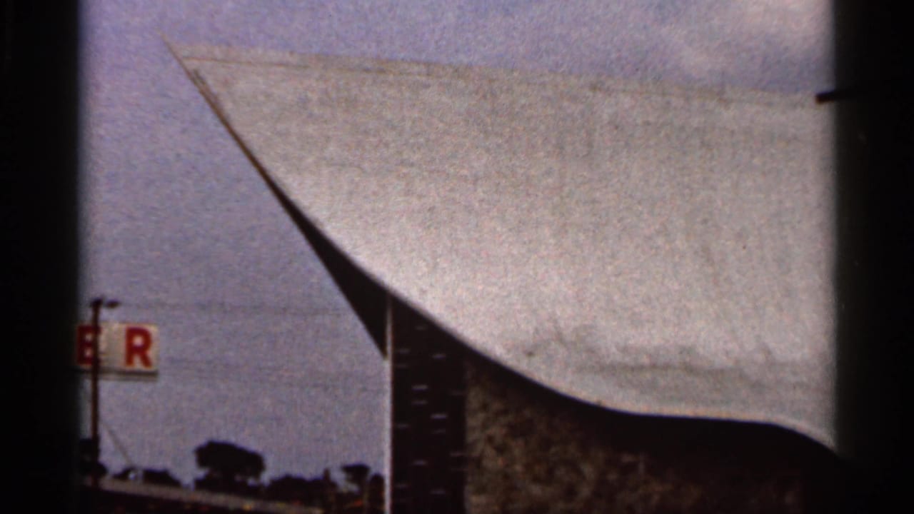 1966 Florida frame of a curved church roof and tall cross, partially obscured by dark vertical edges and a roadside sign.