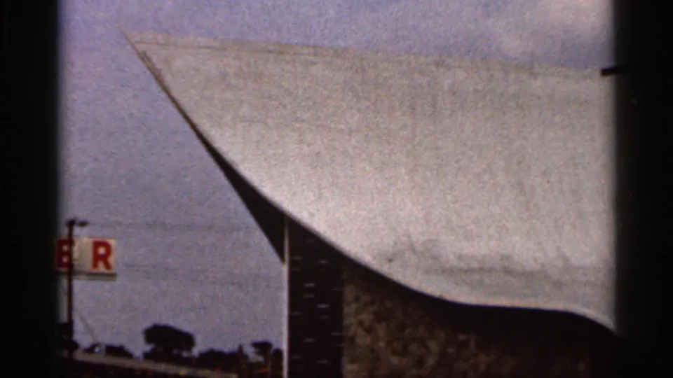 1966 Florida frame of a curved church roof and tall cross, partially obscured by dark vertical edges and a roadside sign.