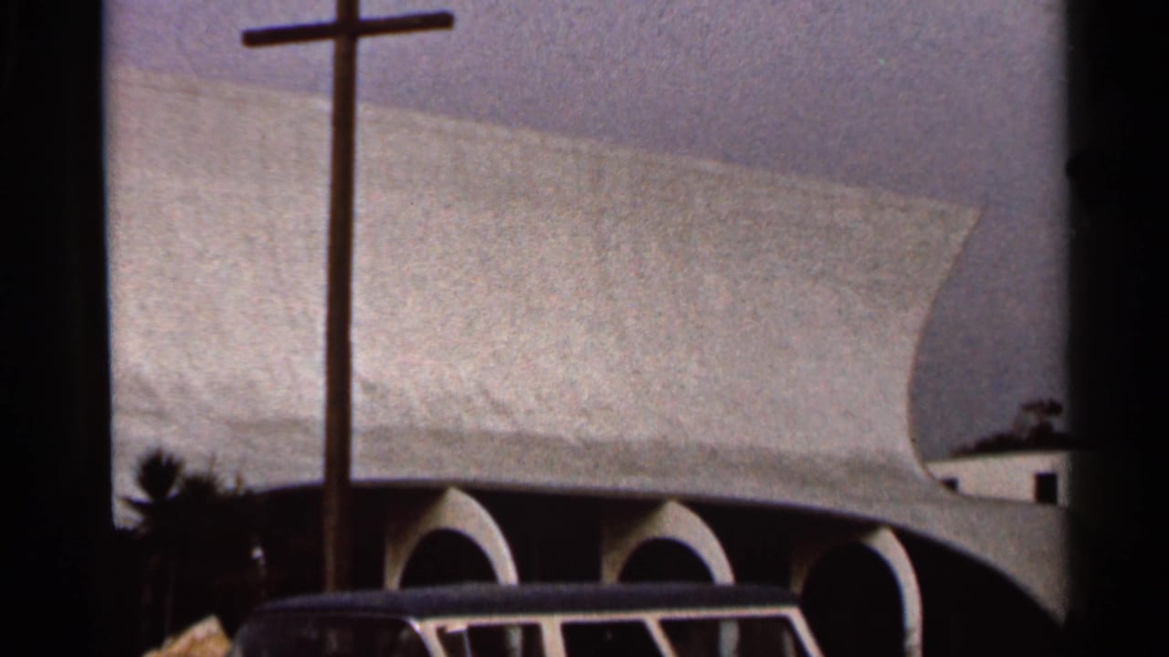 Modern church exterior with tall cross, curved roof, arches, and partial vehicle at the bottom edge.