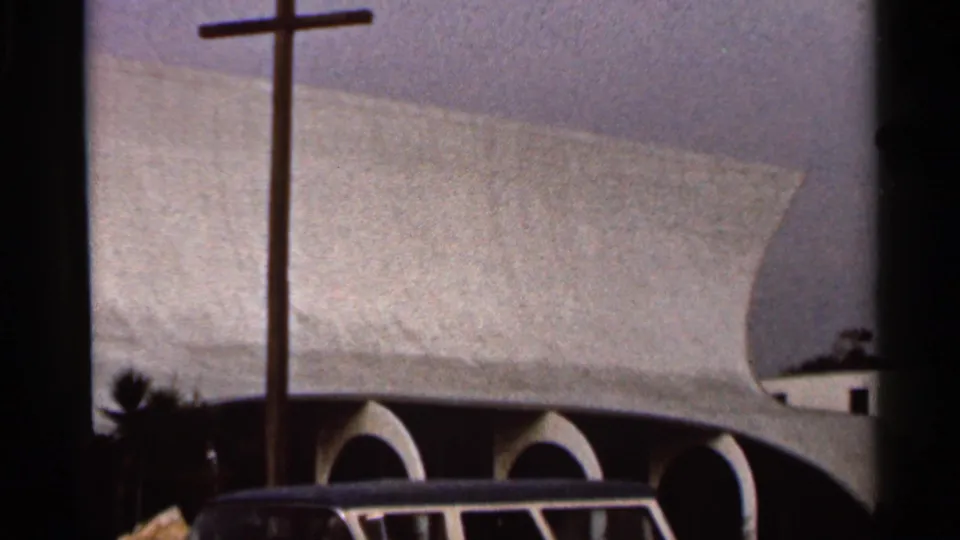 1966 Florida home-movie frame showing a modern church exterior with a tall cross, curved roofline, arched entryways, and a partial vehicle.