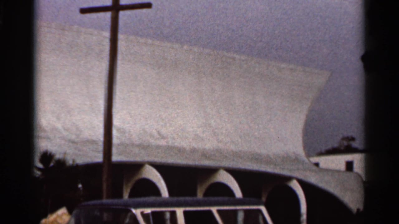 1966 Florida home-movie frame of a church facade with a freestanding cross, sweeping roof, arched bays, and dark 8mm frame edges.
