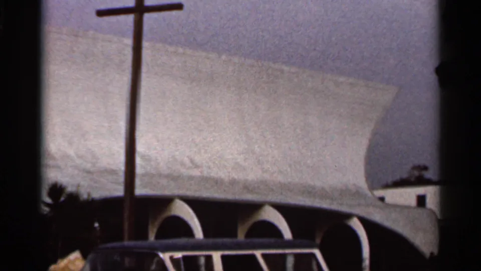 1966 Florida home-movie frame of a church facade with a freestanding cross, sweeping roof, arched bays, and dark 8mm frame edges.