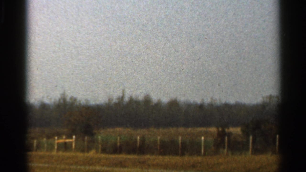 1966 Florida home-movie frame with a low wooden fence, brown field, pale sky, and distant trees framed by dark gate edges.