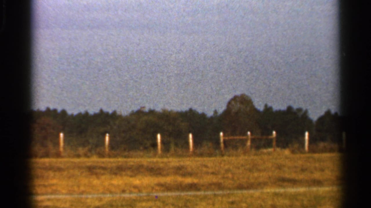 1966 Florida home-movie frame of an open rural field with white fence posts, a wooden rail section, trees, and faded 8mm color.