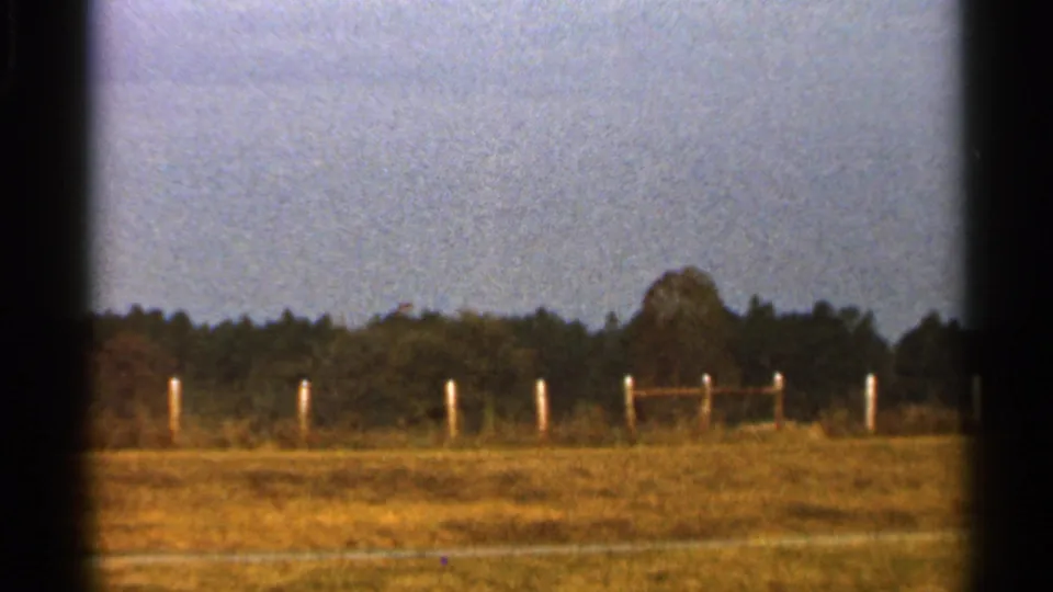 1966 Florida home-movie frame of an open rural field with white fence posts, a wooden rail section, trees, and faded 8mm color.