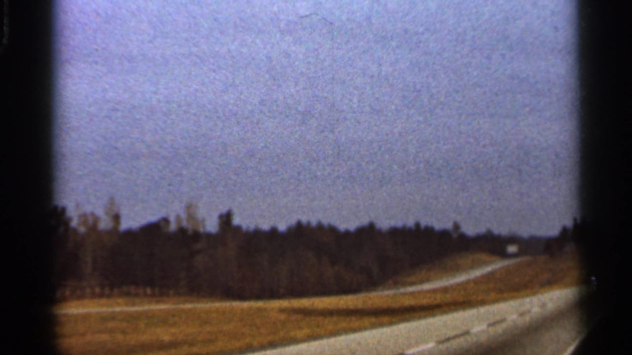 Rural road curving past dry grass and a dark tree line under a hazy sky.