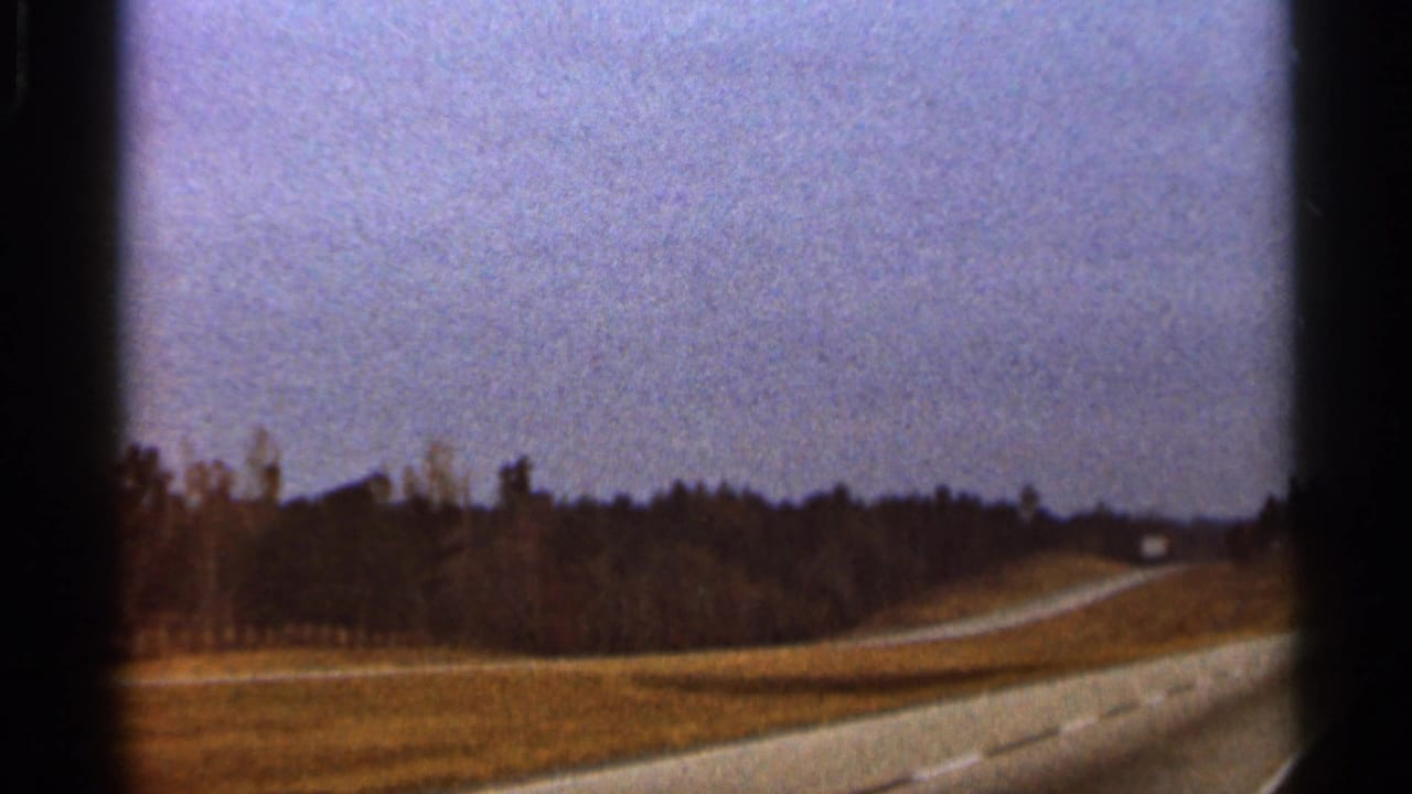 Roadside landscape with open grass, trees, and a curving road seen through dark film edges.