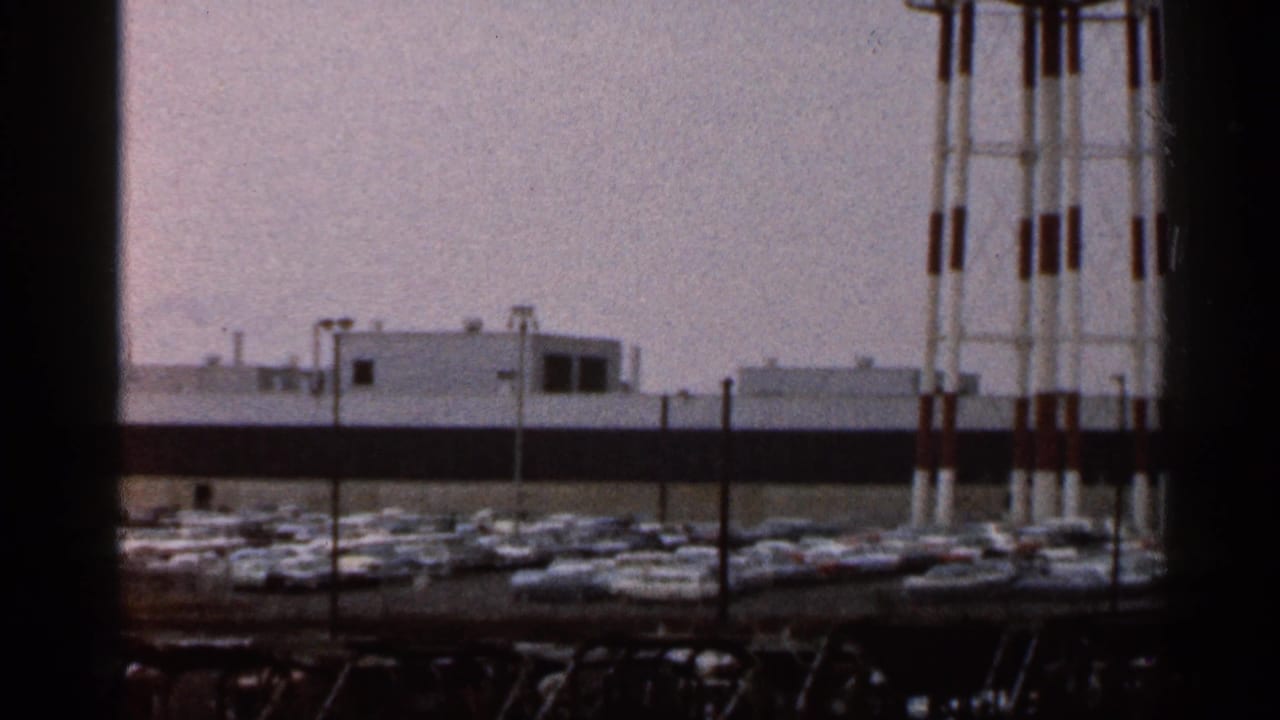 1966 Florida 8mm frame showing low buildings, parked cars, fencing, and tall utility or structural posts near a roadside area.