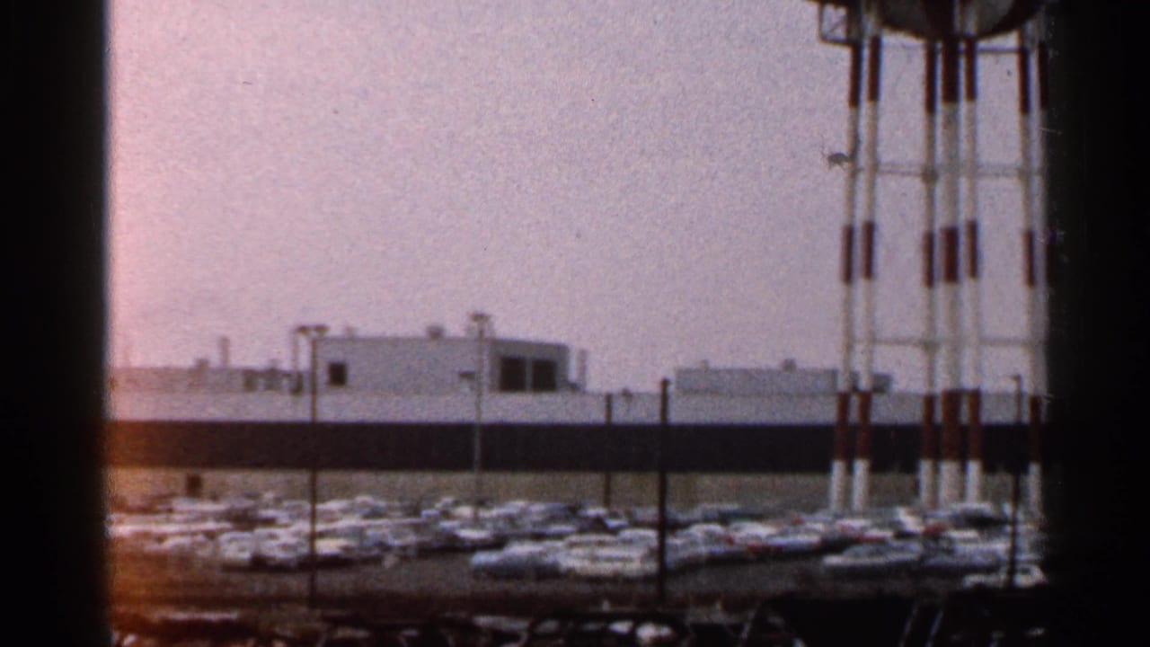 Parked cars fill a lot below a striped water tower and low industrial building.